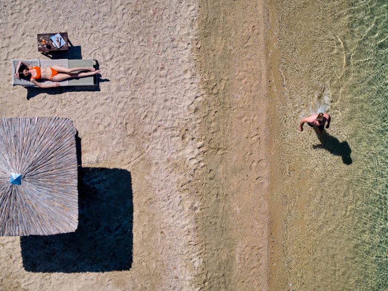 Aerial beach scene: woman in an orange bikini sunbathing on a lounge under a straw umbrella, with a person wading in the turquoise water nearby.