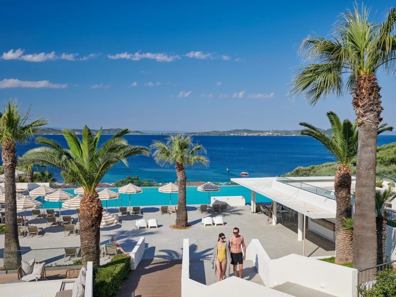 Resort pool area with lounge chairs, striped umbrellas, and tall palm trees overlooking the blue sea.