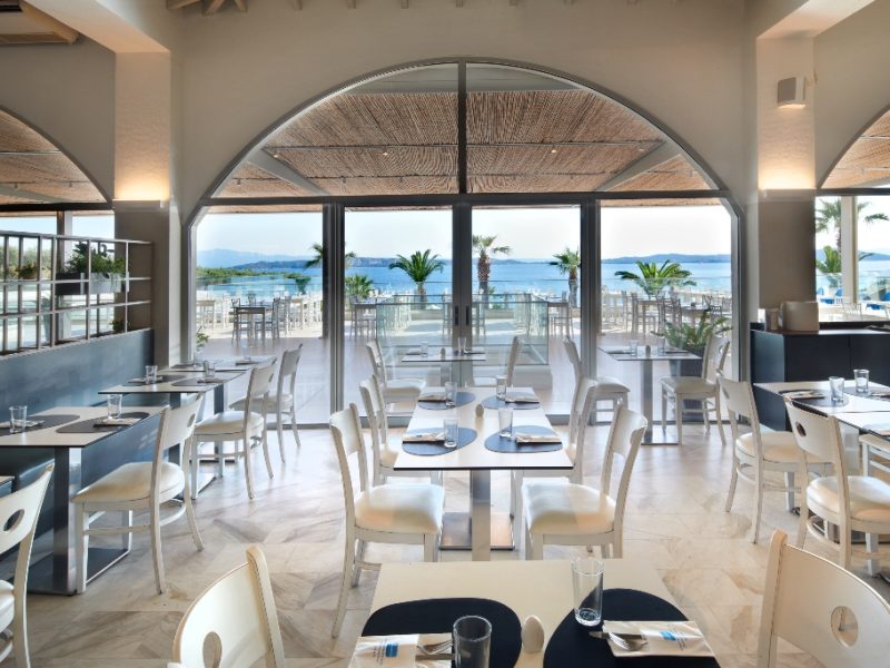 Bright coastal restaurant interior with white tables and chairs, large glass doors opening to a beach and ocean view.
